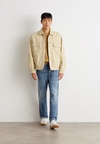 Chaqueta de mezclilla beige con dos bolsillos en el pecho, un suéter beige, jeans azul claro y zapatillas blancas, de pie contra un fondo sencillo.