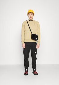 Joven con un gorro amarillo, sudadera beige, pantalones cargo negros, zapatillas rojas y negras, y una mochila cruzada negra, contra un fondo blanco.
