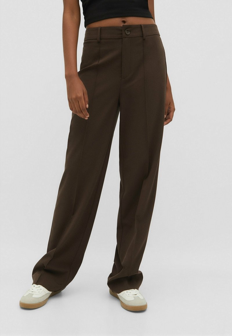 Personne portant un pantalon marron à jambes larges et des baskets blanches, debout devant un fond clair uni.