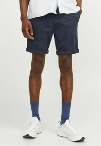 Pantalones cortos azul marino con lunares blancos, dobladillos enrollados y bolsillos laterales. Llevados con calcetines azul y zapatillas deportivas blancas. Textura de tela ligera.