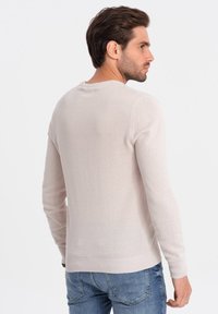 Pull en tricot beige clair à manches longues, col rond, motif texturé subtil, et poignets et ourlet côtelés, associé à un jean bleu.