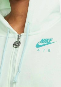 Miętowa zielona bluza Nike z kapturem, wyposażona w srebrny zamek i jasnoniebieskie sznurki. Bluza prezentuje turkusowe logo Nike Air.