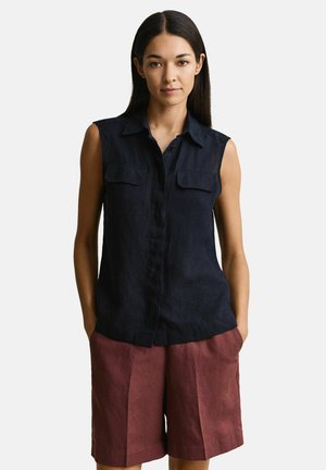 Vrouw die een mouwloze zwarte blouse met knopen draagt, met twee borstzakken en losse bordeauxrode korte broek, staand met handen in de zakken.