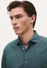 Cămașă button-up teal cu guler, având un logo subtil pe pieptul stâng și detalii cu nasturi. Textură de stofă netedă.