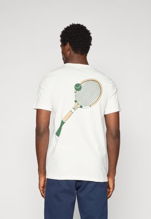 Mand iført en hvid t-shirt med en stor grøn og beige tennisracket og bold grafik på ryggen, kombineret med navy blå shorts.