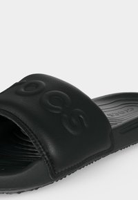 Μαύρο σανδάλι slip-on με υφή πάτου και φαρδιά λουρίδα με χαραγμένο όνομα μάρκας, γωνιακά για να δείξει πλευρικές και ανώτερες λεπτομέρειες.