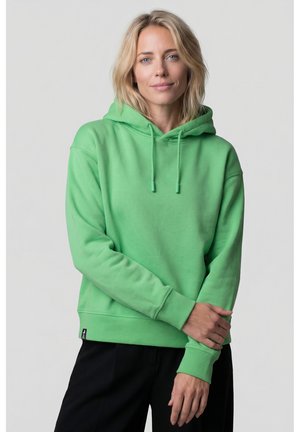 Frau mit blondem Haar, die einen grünen Hoodie und schwarze Hose trägt, steht mit locker verschränkten Armen vor einem schlichten Hintergrund.