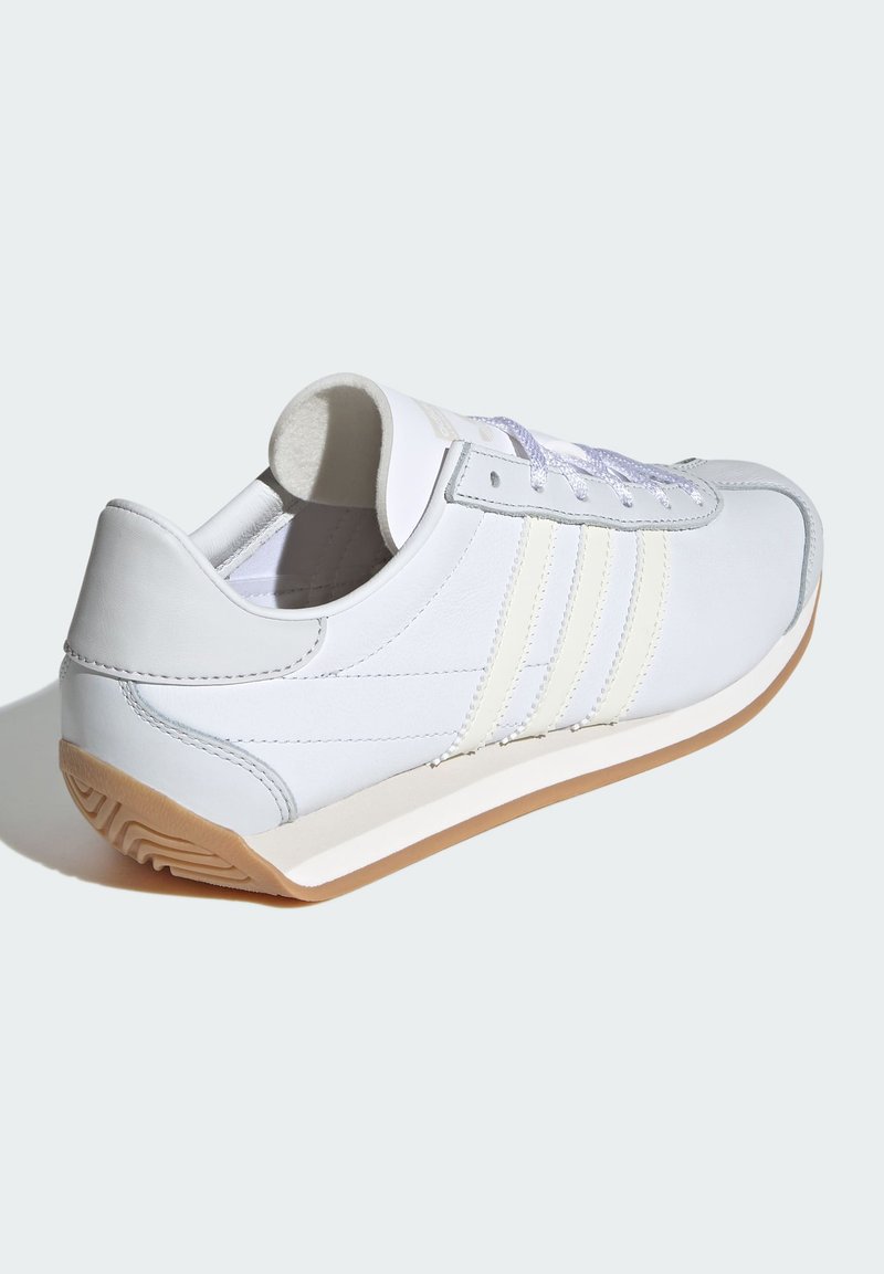 Zapatilla deportiva blanca con parte superior de cuero suave, tres rayas gris claro y suela de goma. Cuenta con cordones planos y un cuello acolchado.