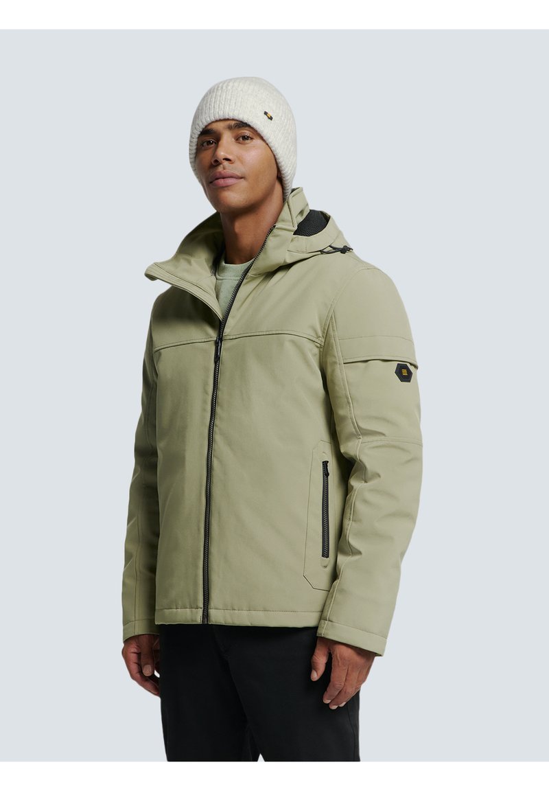 Homme portant une veste zippée vert clair avec des détails de fermeture éclair noirs et un bonnet en tricot blanc, se tenant devant un fond uni clair.