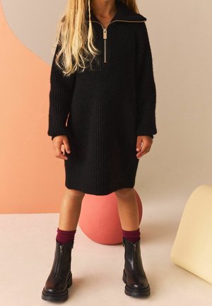 Robe noire en tricot avec un col zippé, manches longues, coupe décontractée. Associée à des bottes noires à fermeture éclair et des chaussettes côtelées bordeaux.