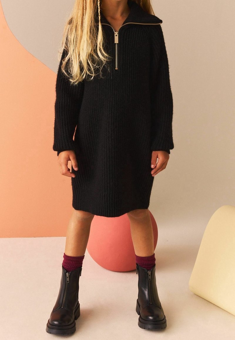 Robe noire en tricot avec un col zippé, manches longues, coupe décontractée. Associée à des bottes noires à fermeture éclair et des chaussettes côtelées bordeaux.