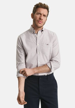 Homme retroussant les manches d'une chemise à rayures claires boutonnée, portée avec un pantalon sombre, sur un fond blanc uni.