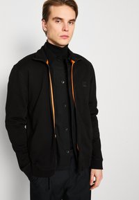 Black zip-up jacket featuring an orange zip detail, soft fabric, and a relaxed fit. Worn over a black button-up shirt.
