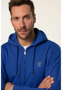 Felpa blu con zip, dotata di corde e logo sul petto, realizzata in materiale morbido, con un design casual.