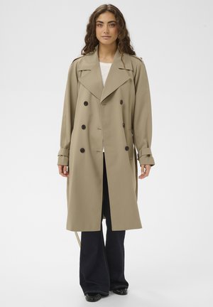 Trench-coat beige croisé avec grands boutons noirs, coupe ajustée, larges revers et manches longues, porté sur un pantalon en denim foncé.