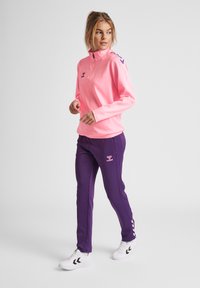 Felpa zip-up rosa chiaro con maniche lunghe e pantaloni viola con logo, entrambi in tessuto liscio. Scarpa sportiva bianca con dettagli neri.