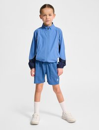 Blå idrottsjacka med marinblå detaljer, dragkedja och ljusgråa kanter; matchande shorts. Vita strumpor och beige skor fulländar outfiten.