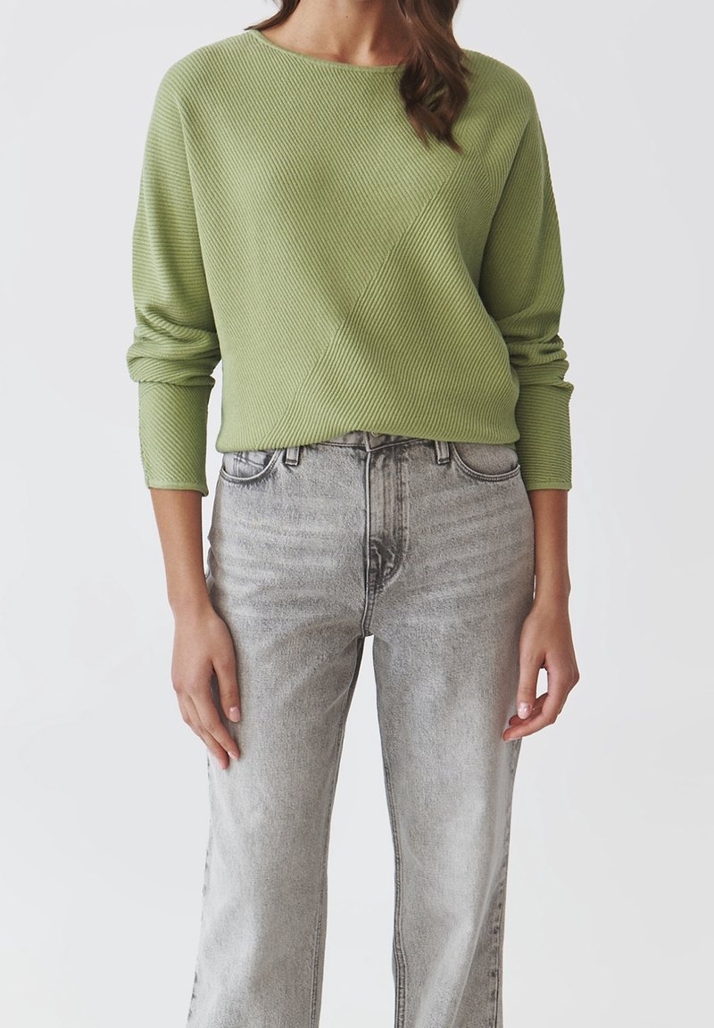 Femme portant un pull en maille côtelée vert clair rentré dans un jean gris délavé taille haute, debout devant un fond uni.