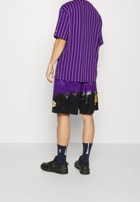 Una persona está de espaldas al espectador, vistiendo una camiseta de rayas moradas, pantalones cortos de teñido anudado en morado y negro, y zapatillas negras.