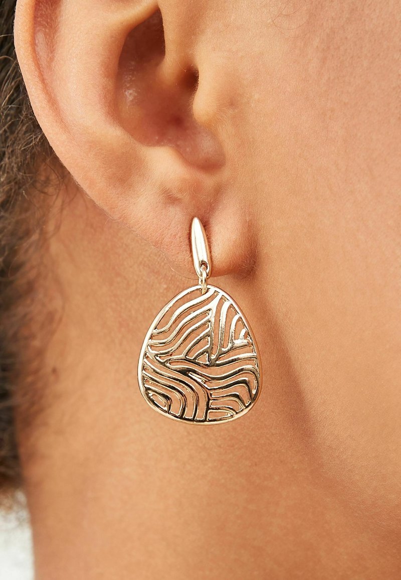 Gros plan d’une boucle d’oreille goutte dorée à motif abstrait portée à l’oreille gauche d’une personne avec un ton de peau neutre.