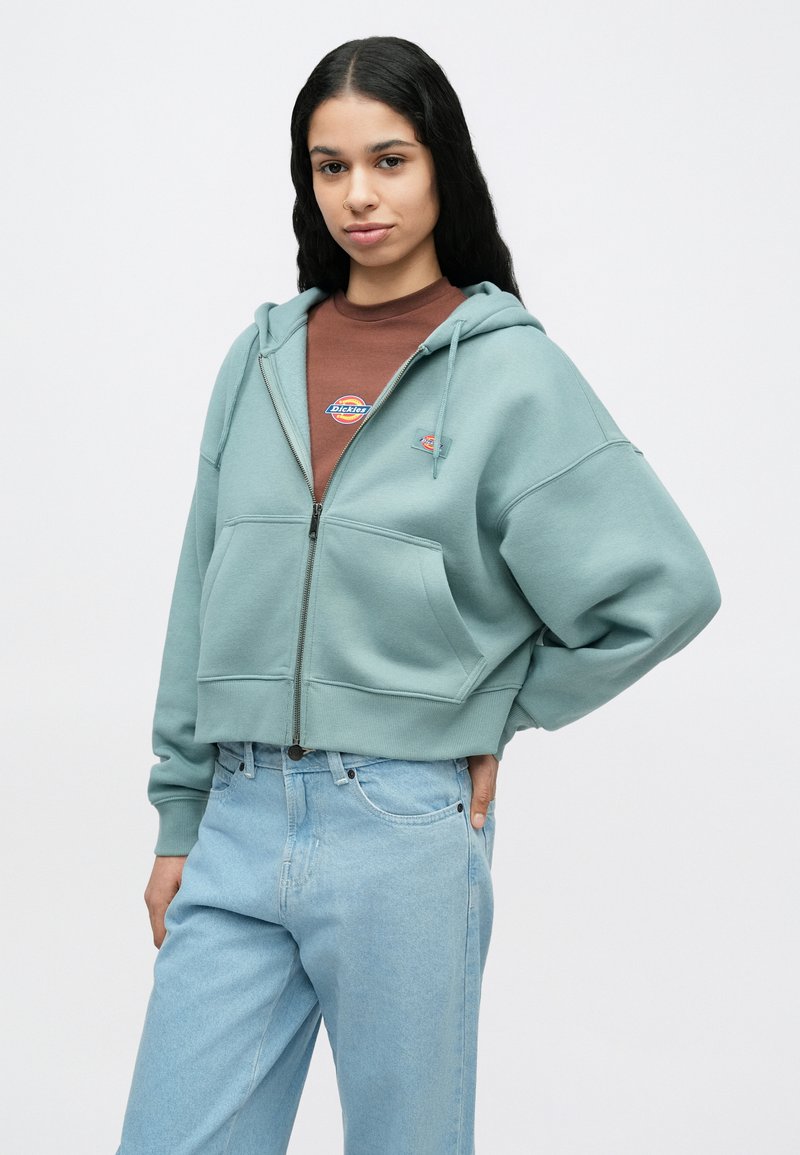 Vrouw met lang donker haar, gekleed in een lichtblauwe hoodie met rits over een bruin T-shirt met logo en lichtblauwe spijkerbroek, poserend met hand op heup.