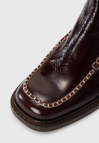 Bottes en cuir bordeaux foncé avec un orteil carré, des coutures beiges contrastantes le long de la couture, et une surface texturée.