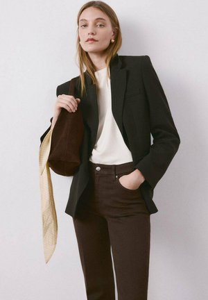 Mujer con blazer negro, blusa blanca, pantalones marrones, pendientes dorados, llevando un bolso marrón con bufanda beige sobre un fondo claro liso.