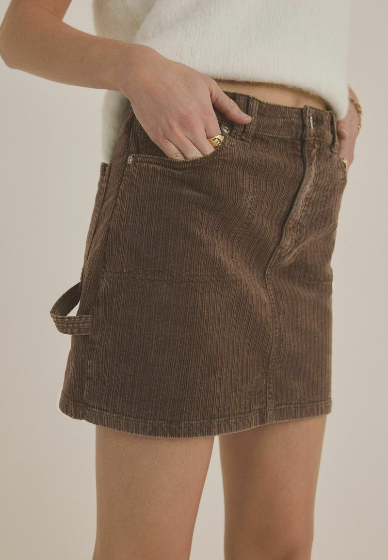 Brun corduroy nederdel med vertikale ribber, sidelommer og bæltestropper. Stoffet har en blød tekstur og en let falmet udseende.