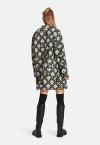 Gemusterter Mantel in Hellgrün mit hexagonalem und floralem Design; lange Ärmel, lockere Passform, kombiniert mit kniehohen schwarzen Stiefeln.