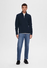 Marinblå zip-up tröja i strukturerad stickad tyg, kombinerad med ljusblå raka jeans och svarta snörskor.
