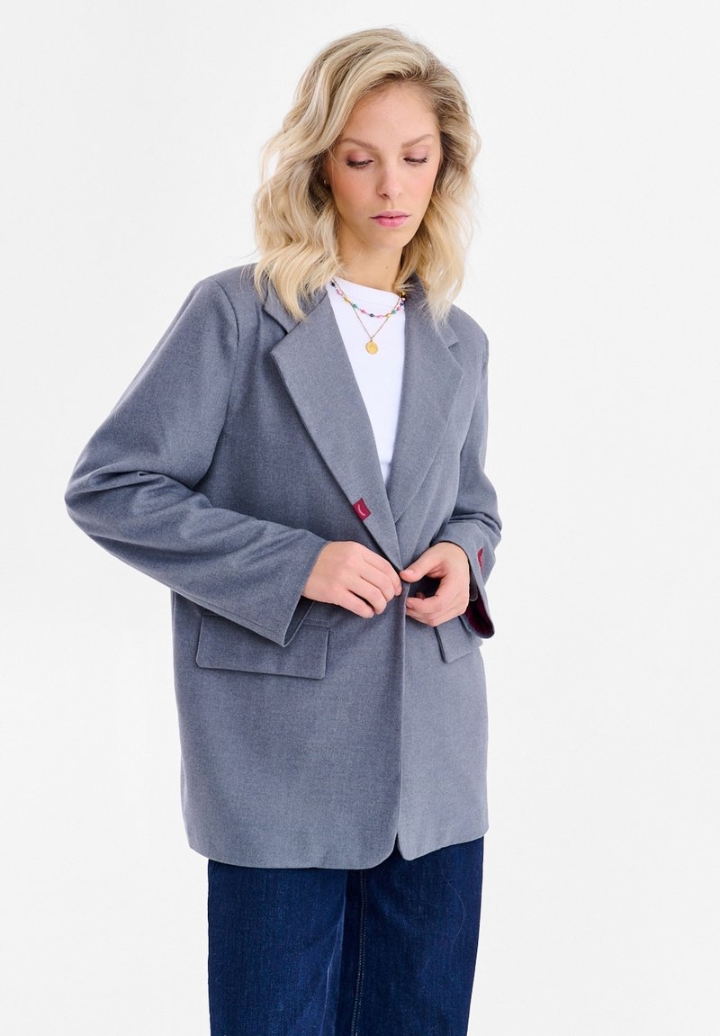 Sivý vlnený blejzer so širokými klopami, jedným červeným gombíkom a vreckami vpredu, nosený cez bielu košeľu a skombinovaný s tmavými džínsovými nohavicami.