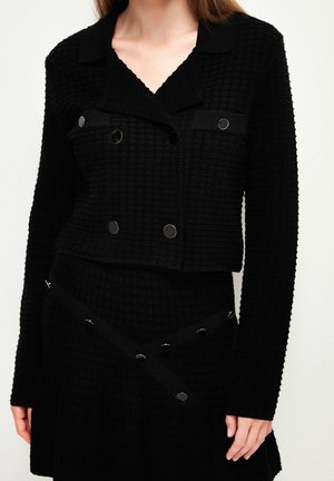 Frau trägt eine strukturierte schwarze Jacke mit langen Ärmeln und metallischen Knopfdetails sowie einen passenden Rock mit diagonal verlaufenden Riemenakzenten.