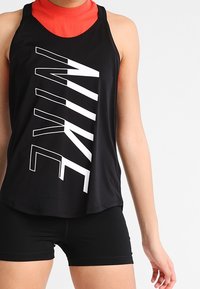 Schwarzes Sport-Tanktop mit großem weißen "NIKE"-Logo, ärmelloses Design, glattes Material, getragen über einem roten Oberteil mit hohem Kragen, kombiniert mit schwarzen Shorts.