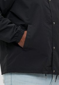 Crna lagana jakna s gumbima, elastičnim rukavima i bočnim džepovima. Glatka tekstura s ravnim rubom.