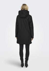 Femme debout de dos portant un manteau à capuche noir, un pantalon ajusté foncé et des bottines à talons noires, sur un fond clair uni.