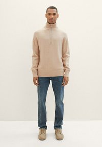 Suéter de punto beige con cuello alto y media cremallera, combinado con jeans azules y zapatos beige, de pie frente a un fondo neutro.