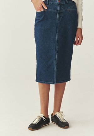 Moteris dėvi tamsiai mėlyną džinsinę kelio ilgio sijoną su priekiniu skeltuku, baltą megztinį ir juodus sportbačius su baltais raišteliais, stovėdama priešais vienodo fono sieną.