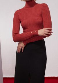 Czerwony sweter z golfem wykonany na drutach, z prążkowaną fakturą oraz małymi złotymi guzikami na mankietach, w połączeniu z czarną spódnicą z rozcięciem.