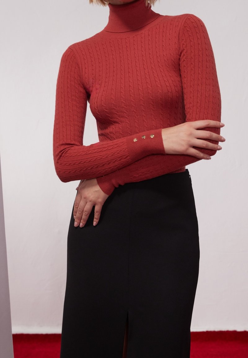 Czerwony sweter z golfem wykonany na drutach, z prążkowaną fakturą oraz małymi złotymi guzikami na mankietach, w połączeniu z czarną spódnicą z rozcięciem.