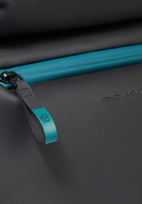 Bolso negro con superficie texturizada, con una cremallera turquesa y una lengüeta a juego. El nombre de la marca "Piquadro" está grabado cerca.