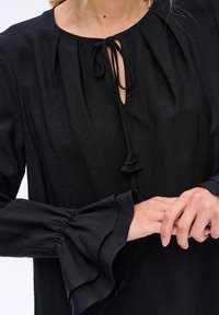 Czarna lniana bluzka z okrągłym dekoltem, z zapięciem na sznurek i falbaniastymi rękawami. Gładka faktura o minimalistycznym designie i bez widocznych wzorów.