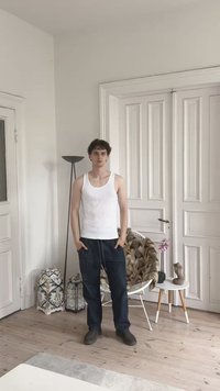 Αρσενικό άτομο ντυμένο με λευκή αμάνικη μπλούζα και σκούρα παντελόνια, όρθιο σε εσωτερικό χώρο, με τα χέρια στις τσέπες του. Ο χώρος διαθέτει ξύλινο πάτωμα και διάφορα διακοσμητικά αντικείμενα.