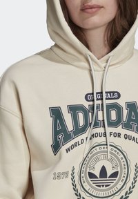 Primo piano di una persona che indossa un hoodie crema Adidas con logo verde, con il testo "WORLD FAMOUS FOR QUALITY" e i laccetti visibili nell'area del petto.