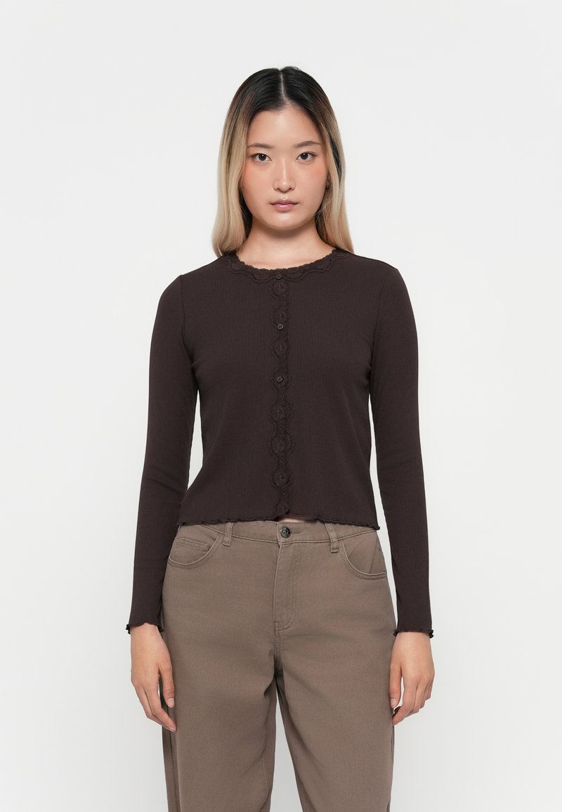 Brown ribbed long-sleeve top with a scalloped collar and button closure down the front. Worn with light brown pants.