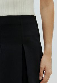 Schwarze Rock mit plissiertem Design, strukturiertem Stoff und unsichtbarem Reißverschluss an der Seite. Nahaufnahme der Taille und einer Hand, die auf dem Stoff ruht.