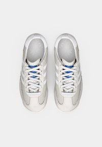 Bézs sportcipő fehér csíkokkal, texturált velúr és bőr felsőrésszel, fűzős kialakítással, párnázott gallérral és kék logóval a nyelén.