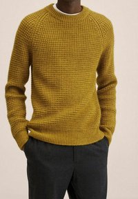 Sweter w musztardowym kolorze, wykonany na drutach, z teksturalnym wzorem, ściągaczem przy dekolcie i u dołu oraz rękawami raglanowymi. Charakteryzuje się luźnym krojem.