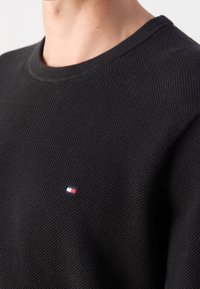 Gros plan sur une personne portant un pull à col rond noir texturé avec un petit logo rectangulaire rouge, blanc et bleu sur la poitrine.