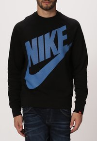 Černá mikina s velkým modrým logem Nike se pruhovaným vzorem, pružnými manžetami a lemem. Vyrobena z měkké tkaniny, vhodná pro volnočasové nošení.