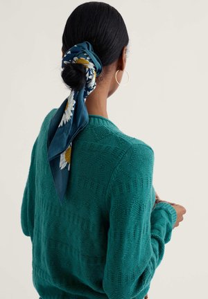 Mujer con cabello oscuro, lleva un suéter texturizado color verde azulado, una bufanda azul con estampado floral atada alrededor de un moño bajo y aretes de aro dorados, vista de espaldas.